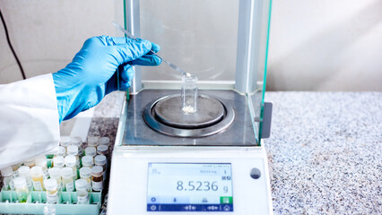 A scientist's gloved hand uses a stainless spatula to transfer a substance into a vial for weighing...