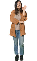Full-body young Chinese woman winks an eye and holds an okay gesture with hand.