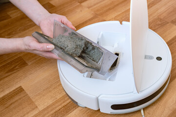 woman removes a container of dust from a round white robot vacuum cleaner. Modern electronics for...