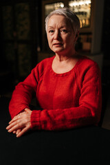 Obraz premium Vertical portrait of attractive gray-haired senior woman in casual red sweater looking at camera with serious expression while sitting at table in dark room, by light of burning candle.