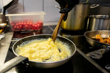 A hand in a black glove stirs scrambled eggs in a frying pan on a stove, with pots and ingredients in the background