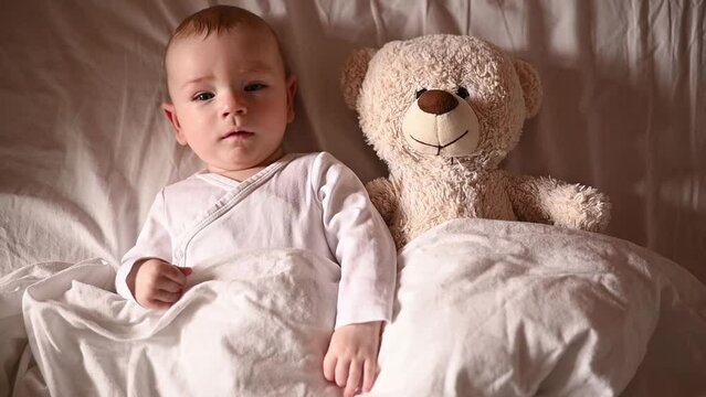 little cute baby lying with teddy bear under blanket on big bed