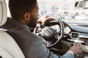 Black businessman in a suit drives a modern car on a sunny day in an urban environment. His hand...