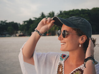 Woman Adjusting Cap and Smiling Brightly on a Scenic Beach Sunset - Concept of Happiness and Adventure by the Sea