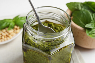 Tasty pesto sauce in glass jar, spoon, pine nuts and basil on table, closeup