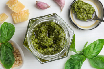 Tasty pesto sauce in jar, spoon, pine nuts, garlic and basil on white wooden table, flat lay
