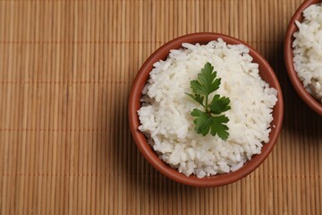 Bowls of delicious boiled rice with parsley on bamboo mat, top view. Space for text
