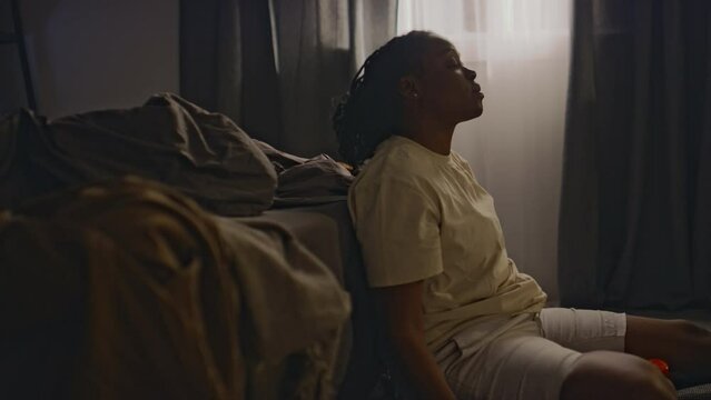Side shot of young Black woman in loungewear sitting on floor by unmade bed and taking tranquilizing drug after sleepless night, living with insomnia