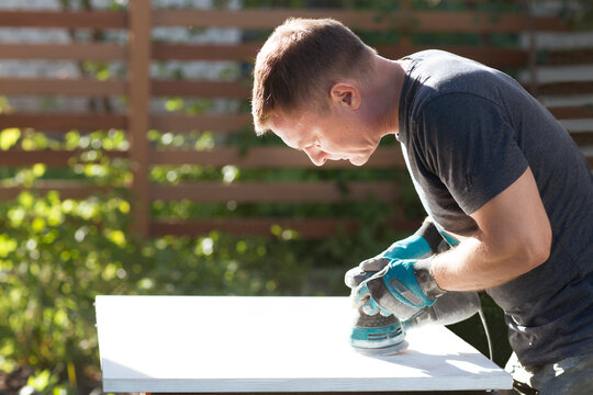the man carpenter  is engaged in steel work near the house in the summer. a man using wood polishing machine grinding sanding wooden board surface on workbench.  master class workshop.