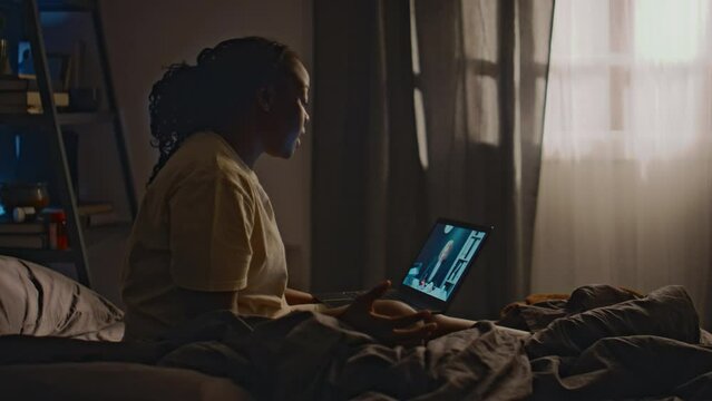 Side shot of young Black woman speaking with psychiatrist during online therapy session on her laptop sitting in unmade bed after sleepless night, discussing ways to cope with insomnia