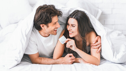 A couple is lying in bed, both smiling, with the woman holding a condom. They are under the covers...