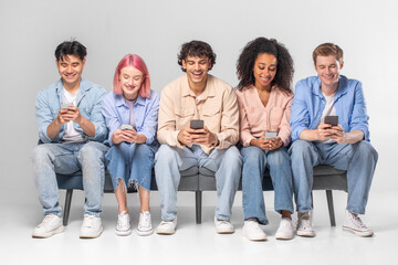 This image shows five diverse young adults sitting on a bench together. They are all looking at their smartphones. The people are casual and relaxed.