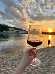 A glass of red wine in a woman's hand during sunset in a big city on a river bank
