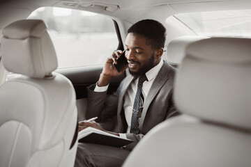 African American businessman in a suit sits in the backseat of a car. He is talking on his phone...