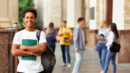 Obraz premium Enjoying university life. African guy holding books and smiling, standing against friends