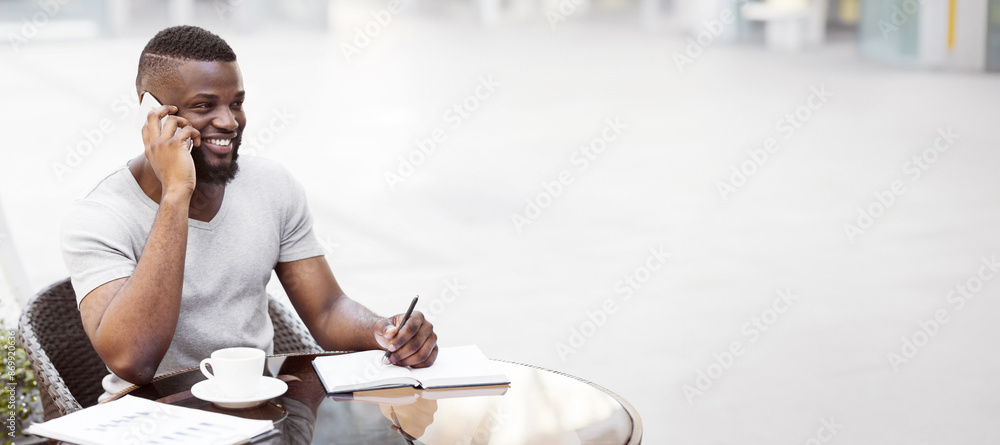 Poster a man sits at a cafe table, talking on his phone and writing in a notebook, smiling while on a call.