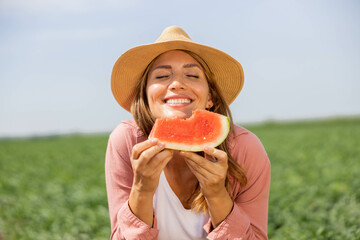 portrait of a beautiful young farmer woman with a hat in a field holding a piece of watermelon. she enjoys eating the ripe watermelon she picked