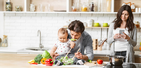 A father feeds his baby a salad while sitting at the kitchen counter. The baby is happily eating the salad, while the father smiles at the baby. The mother stands behind them, copy space