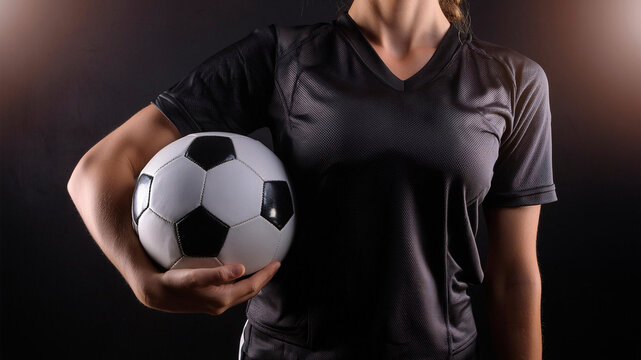 F&uacute;tbol femenino. Mujer futbolista con camiseta negra de deporte sujetando con una mano un bal&oacute;n de f&uacute;tbol. Concepto de igualdad, superaci&oacute;n empoderamiento