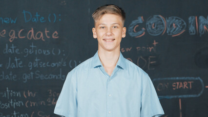 Smiling teenager looking at camera while standing at blackboard with engineering code or prompt...