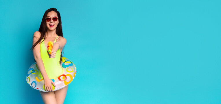 A woman wearing a yellow swimsuit and sunglasses is holding a water gun and a colorful pool float in front of a solid blue background. She is smiling and looking directly at the camera