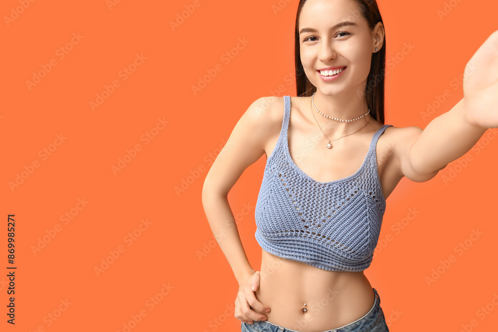 Sticker Beautiful young happy woman with piercing navel taking selfie on orange background