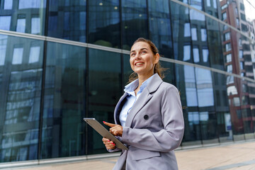 Fototapeta premium A businesswoman in a grey suit holds a tablet and smiles while standing outside a modern glass building.