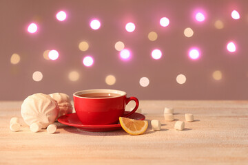 Red cup with hot brewed tea, slices of lemon and marshmallow on white wooden table against blurred background