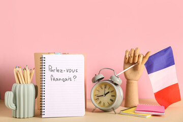 Notebook with French phrase, flag, stationery and alarm clock on table against color background
