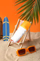 Bottle of sunscreen cream on deckchair, sunglasses and surfboards on sand against orange background