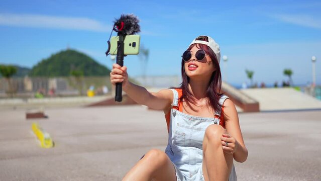 Asian young woman are doing livestreaming joyfully in skateboard urban park waving at the camera while recording