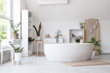 Bathtub, shelving units, chest of drawers and houseplants in modern bathroom