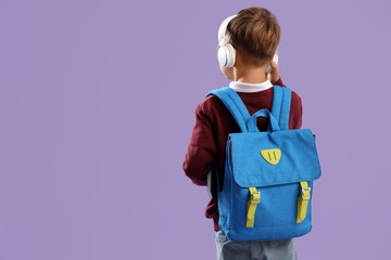 Little schoolboy in headphones with backpack on lilac background, back view