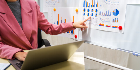 A businesswoman in a pink suit works at a desk with a whiteboard in the background. She focuses on accounting and financial services, professionalism and expertise in various financial roles.