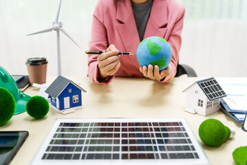 A businesswoman in a pink suit stands at a white desk with a world model, electric car model, and green light bulb, promoting Earth Day, green energy, and sustainability efforts.