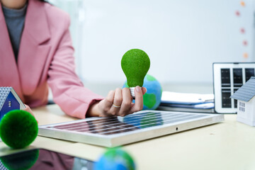 A businesswoman in a pink suit stands at a white desk with a world model, electric car model, and green light bulb, promoting Earth Day, green energy, and sustainability efforts.