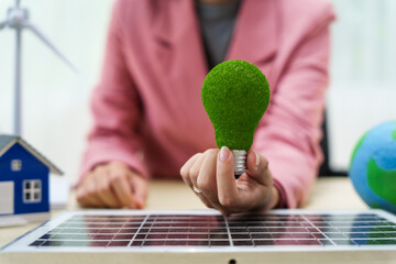 A businesswoman in a pink suit stands at a white desk with a world model, electric car model, and green light bulb, promoting Earth Day, green energy, and sustainability efforts.