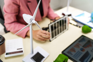 A businesswoman in a pink suit stands at a white desk with a world model, electric car model, and green light bulb, promoting Earth Day, green energy, and sustainability efforts.