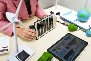 A businesswoman in a pink suit stands at a white desk with a world model, electric car model, and green light bulb, promoting Earth Day, green energy, and sustainability efforts.
