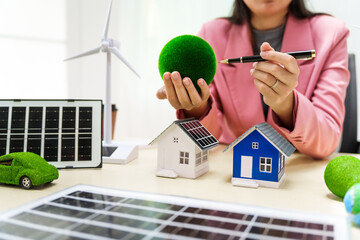 A businesswoman in a pink suit stands at a white desk with a world model, electric car model, and green light bulb, promoting Earth Day, green energy, and sustainability efforts.
