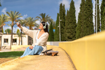 Young Latina woman with headphones, sitting on yellow steps in a park, taking a selfie with her smartphone. background. lifestyle concept