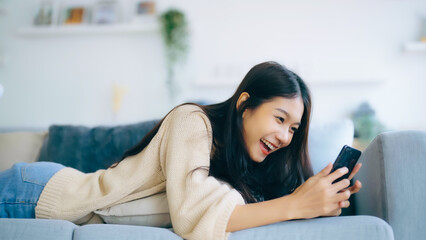 Happy young asian woman relax on comfortable couch at home texting messaging on smartphone, smiling girl use cellphone, browse wireless internet on gadget, shopping online from home