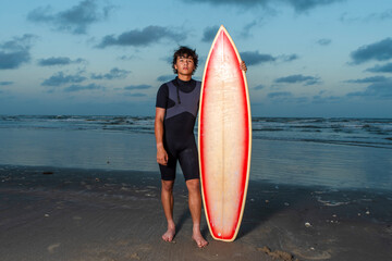 Handsome wet surfer boy standing next to his surfboard after a long day of surfing comes to an end