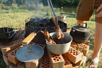 Fototapeta premium A man stands in nature, preparing meat on a makeshift grill built from bricks. He uses tongs to transfer the food from the grill to a pot. Tourist on recreation outside. Campsite lifestyle