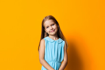 Little girl in a blue dress with hands in pockets stands confidently against a vibrant orange background