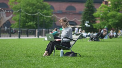 Focused woman with computer sitting on folding chair on lawn in busy public place. Peaceful businesswoman working in open air during day. Calm freelance girl attentively performing project tasks.