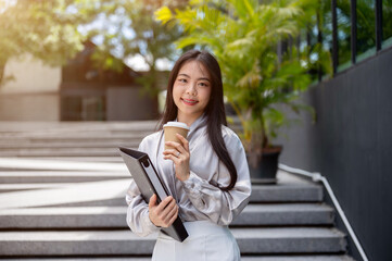 Obraz premium Attractive Asian businesswoman with a file folder and a coffee cup, standing on the stairs outdoors.