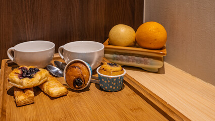 There are two white earthenware cups and teaspoons on a wooden tray. Muffins, sweet pastries are laid out.  There is an orange and an apple on the table. Breakfast is ready.