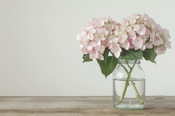 A bouquet of pastel pink hydrangeas in a clear glass jar filled with water, placed on a rustic wooden table against a plain white background.