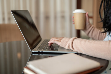 A cropped image of a woman working on her laptop computer and having a hot drink at a table indoors.